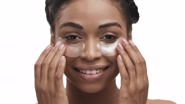 Cheerful young african american lady applying gold anti-fatigue patches under eyes, smiling to camera, white background