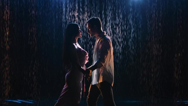 Silhouettes loving couple whirl in dance, closely touching their bodies and holding hands. Romantic dance in dark studio against backdrop of falling raindrops and blue light. Close up. Slow motion.