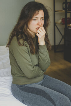 Worried sad young brunette woman thinking about problems, biting nails sitting on bed at home interior. Unhappy teenager suffering from psychological problem, unexpected pregnancy concept. Vertical.