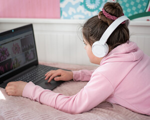 Dziewczynka pracuje zdalnie w domu przed komputerem ze słuchawkami na uszach leżąc na łóżku. - obrazy, fototapety, plakaty