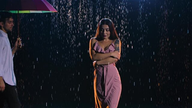Love story of couple in rain, expressed in passionate dance. Romantic meeting of two lovers. Couple under bright colored umbrella. Wet bodies of man and woman in studio light. Close up. Slow motion.