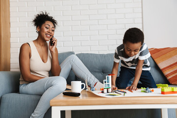 Black smiling woman talking on cellphone while her son playing with toys