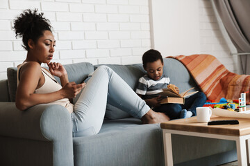 Black woman using cellphone while her son reading book at home