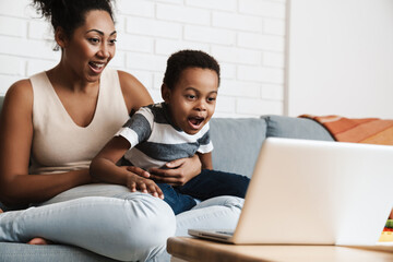Fototapeta premium Black excited mother and son using laptop while sitting on sofa at home