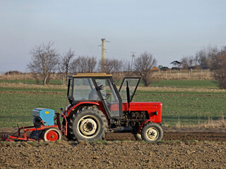 Obraz premium A farmer on a red tractor with a seeder sows grain in plowed land in a private field in the village area. Mechanization of spring field work. Farmer's everyday life. relief of human labor