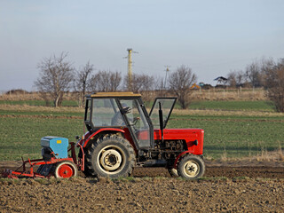 A farmer on a red tractor with a seeder sows grain in plowed land in a private field in the village area. Mechanization of spring field work. Farmer's everyday life. relief of human labor