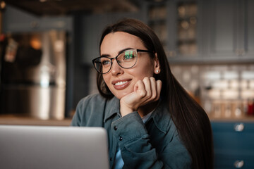 White smiling woman in eyeglasses working with laptop at home kitchen
