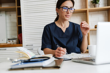 A woman in a medical suit sitting at a table in front of a laptop and looking into it