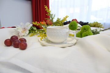 The table is covered with a white airy cloth, served with a white cup on a saucer with red grapes, white, yellow and green flowers
