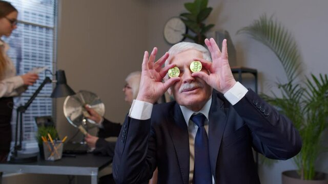 Elderly man boss dancing, fooling around, making silly faces with bitcoin coints eyes in modern office room interior. Senior entrepreneur freelancer celebrating income from stock market or mining