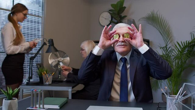 Funny business man entrepreneur dancing, fooling around, making silly faces with bitcoin coints. Senior male professional celebrating income from stock market or mining at office desk. Coworking