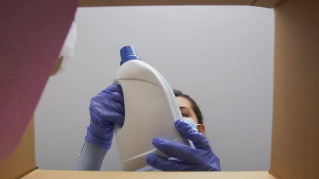 Home Delivery, Shipping And Pandemic Concept - Woman In Protective Medical Mask And Gloves Opening Parcel Box, Looking Inside And Taking Cleaning Supplies Out