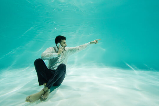 Smiling muslim businessman talking on smartphone and pointing with finger near bottom of swimming pool - Powered by Adobe