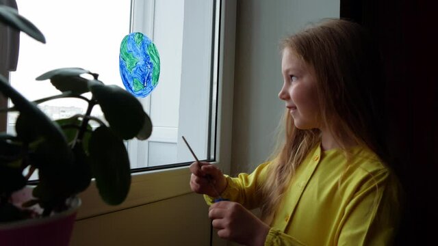 Cute little girl painting planet on window at home 4K. Happy Earth Day April 22 greeting message. Creative family leisure lockdown new reality. Ecology Saving environment conscious consumption concept