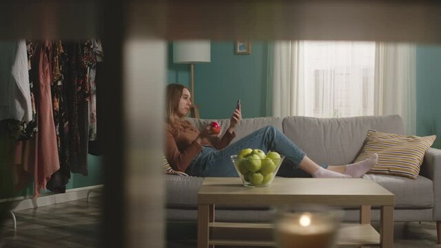 Beautiful girl in blue jeans and brown sweater lies on beige sofa on background of window, biting a red apple and and speaks via video link.