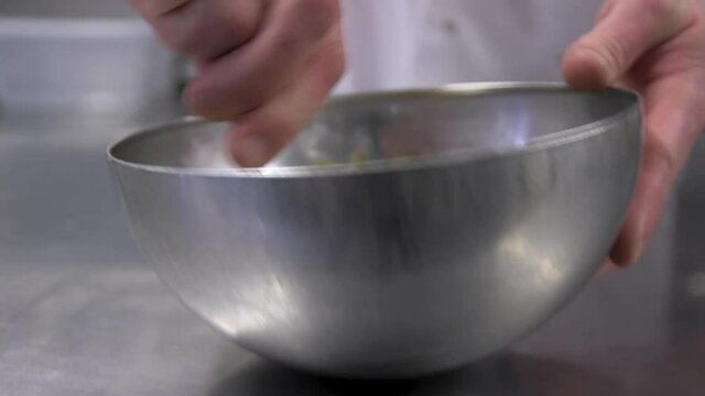 A male professional chef's hands mixing ingredients together by hand using and spoon in a stainless steel bowl in the kitchen of a hotel or restaurant.