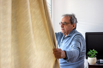 Fototapeta premium Elderly man staying home for safety during coronavirus pandemic and observing empty city through the window. Man staying home during coronavirus pandemic is looking through the window.