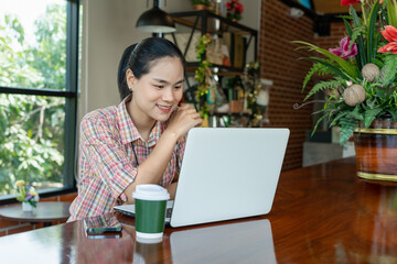 Fototapeta premium Cheerful young Asian woman using laptop while sitting on chair alone at coffee shop. Attractive female smiling with good news or chatting online in social network. Positive and Lifestyle concept