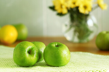 green fresh apples in the kitchen, just washed lie on a towel dry, water drops on the fruit, in the background lemon and a vase with yellow flowers blurred