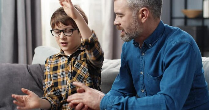 Loving father sits on couch at home and communicates with his school-age son, listening intently to stories.
