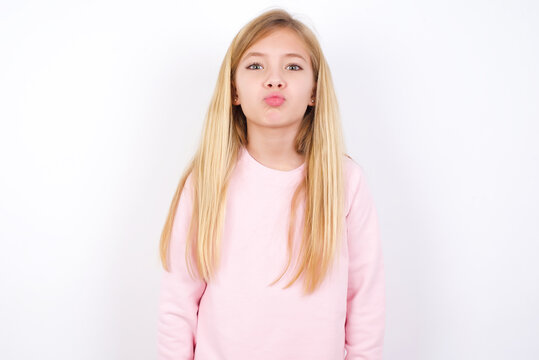 beautiful caucasian little girl wearing pink hoodie over white background, keeps lips as going to kiss someone, has glad expression, grimace face. Standing indoors. Beauty concept.