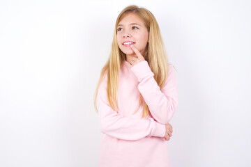 beautiful caucasian little girl wearing pink hoodie over white background with thoughtful expression, looks to the camera, keeps hand near face, bitting a finger thinks about something pleasant.