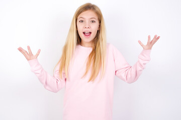 Fototapeta na wymiar Joyful excited lucky beautiful caucasian little girl wearing pink hoodie over white background cheering, celebrating success, screaming yes with clenched fists