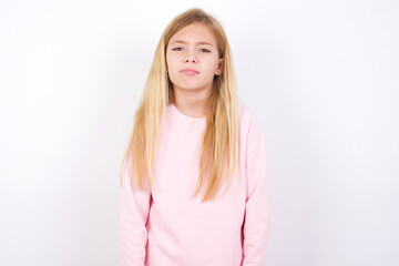 Gloomy, bored beautiful caucasian little girl wearing pink hoodie over white background frowns face looking up, being upset with so much talking hands down, feels tired and wants to leave.