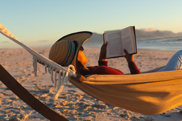 Mixed race woman on beach holiday lying in hammock reading book during sunset