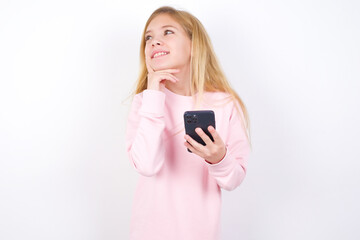 Image of a thinking dreaming beautiful caucasian little girl wearing pink hoodie over white background using mobile phone and holding hand on face. Taking decisions and social media concept.