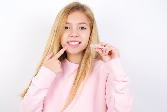 beautiful caucasian little girl wearing pink hoodie over white background holding an invisible aligner and pointing to her perfect straight teeth. Dental healthcare and confidence concept.