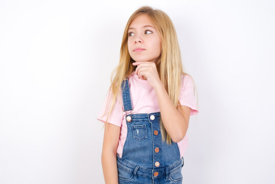 Thoughtful beautiful Caucasian little girl wearing denim overalls over white background holds chin and looks away pensively makes up great plan