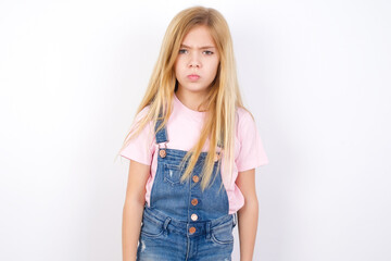 Offended dissatisfied beautiful Caucasian little girl wearing denim overalls over white background with moody displeased expression at camera being disappointed by something
