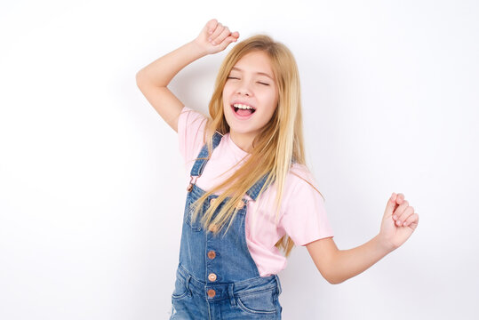 Photo of upbeat beautiful Caucasian little girl wearing denim overalls over white background has fun and dances carefree wear being in perfect mood makes movements. Spends free time on disco party