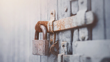 An old metal large lock hangs on a vintage wooden gray gate with a rusty latch, illuminated by light. Closed.