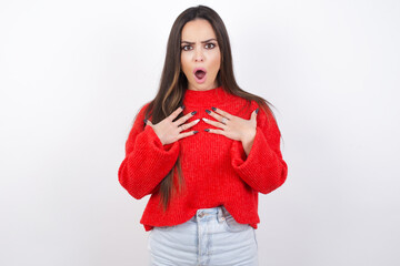 young beautiful brunette woman wearing red knitted sweater over white wall keeps hands on chest feeling shocked and scared, mouth widely opened, stares at camera saying: Who, me?