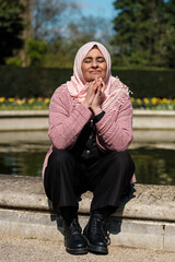 Muslim mature woman wearing hijab enjoying sun in a beautiful spring day. Sitting on the edge of a fountain in a London Park.