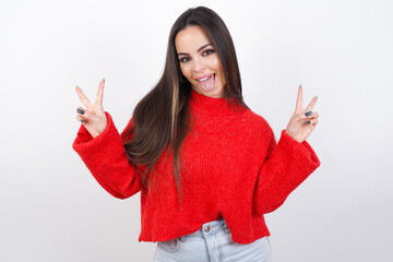 young beautiful brunette woman wearing red knitted sweater over white wall with optimistic smile, showing peace or victory gesture with both hands, looking friendly. V sign.