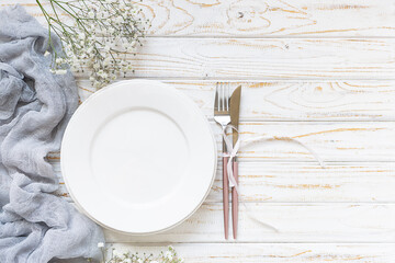 Festive table setting concept. White empty plate, cutlery set, napkin and beautiful flowers on white wooden table top view.