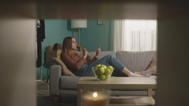 Beautiful woman in blue jeans and brown sweater lies on beige sofa on background of window, biting a red apple and and speaks via video link.