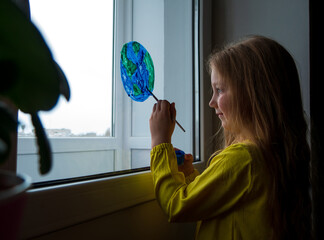 Cute little girl painting planet on window at home. Happy Earth Day April 22 greeting message. Creative family leisure lockdown new reality. Ecology Saving environment conscious consumption concept.