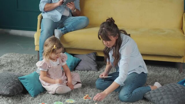 Mom is always there to help. Little girl playing with mother on the carpet, while her father works using a tablet. Spending time together at home.