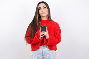 Portrait of young beautiful brunette woman wearing red knitted sweater over white wall with dreamy look, thinking while holding smartphone. Tries to write up a message.