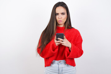 Portrait of a confused young beautiful brunette woman wearing red knitted sweater over white wall holding mobile phone and shrugging shoulders and frowning face.