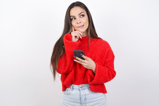Image of a thinking dreaming young beautiful brunette woman wearing red knitted sweater over white wall using mobile phone and holding hand on face. Taking decisions and social media concept.