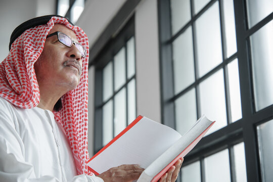 Close up of Elderly Arabian Muslim man in traditional clothes holding and reading holy Quran book. Side view