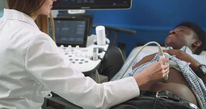 Cropped close up image of ultrasound scanner device in the hand of a professional female doctor examining young African man patient, doing abdominal ultrasound scanning sonography