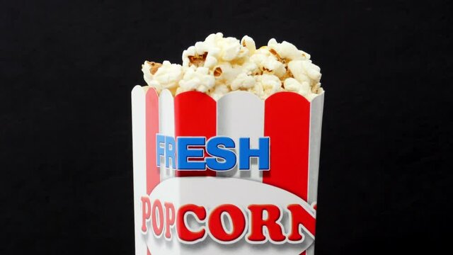 Paper red-white box full of popcorn revolving on black background words fresh popcorn written on it movie time going to cinema entertainment food free time. Close up 4K Ultra HD.