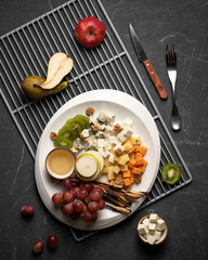 Plate with Cheese Assortment on black table with metal grill and cutlery on it. Top view. Gourmet snack from fruits, berries, nuts and mixed of cheeses.