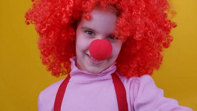 Funny clown girl kid with a red nose and hair is jumps and laughing on a yellow background. 1 April Fool's day, Birthday concept.